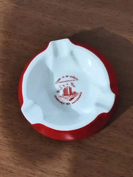Foto de um(a) Cinzeiro Porcelana Steatita, Década de 1960, da categoria de Cinzeiros e coletores. Cinzeiro de mesa em porcelana branca com moldura em vermelho e o logo do clube 12 de agosto, fundado em 12 de agosto de 1872... Mostrado de uma perspectiva diferente. Vendido no marketplace Trésors.
