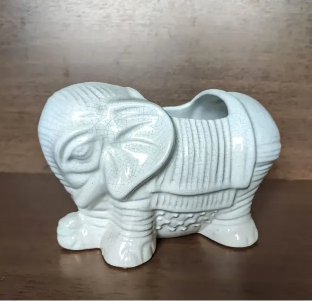 Foto de um(a) Cachepô Elefante Anos 90, da categoria de Cachepot. Cachepô formato de elefante em porcelana craquelada na cor branca, década de 1990. A: 12,5 cm / L: 20cm / P: 12 cmPeso: 704g. Mostrado de uma perspectiva diferente. Vendido no marketplace Trésors.